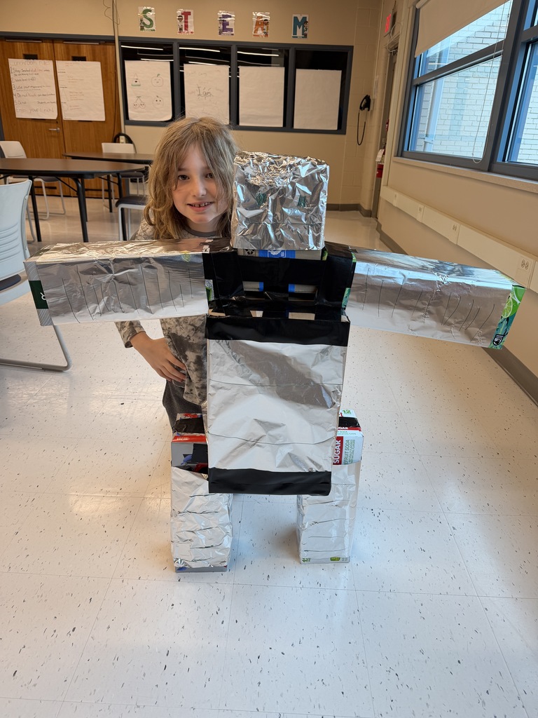 A smiling student kneels behind their robot constructed of boxes, black tape, and tin foil