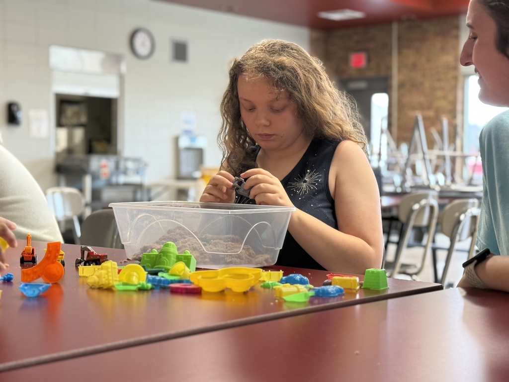 A student sits at a table using their hands to explore sensory materials in a bin filled with textured items and small toys. 