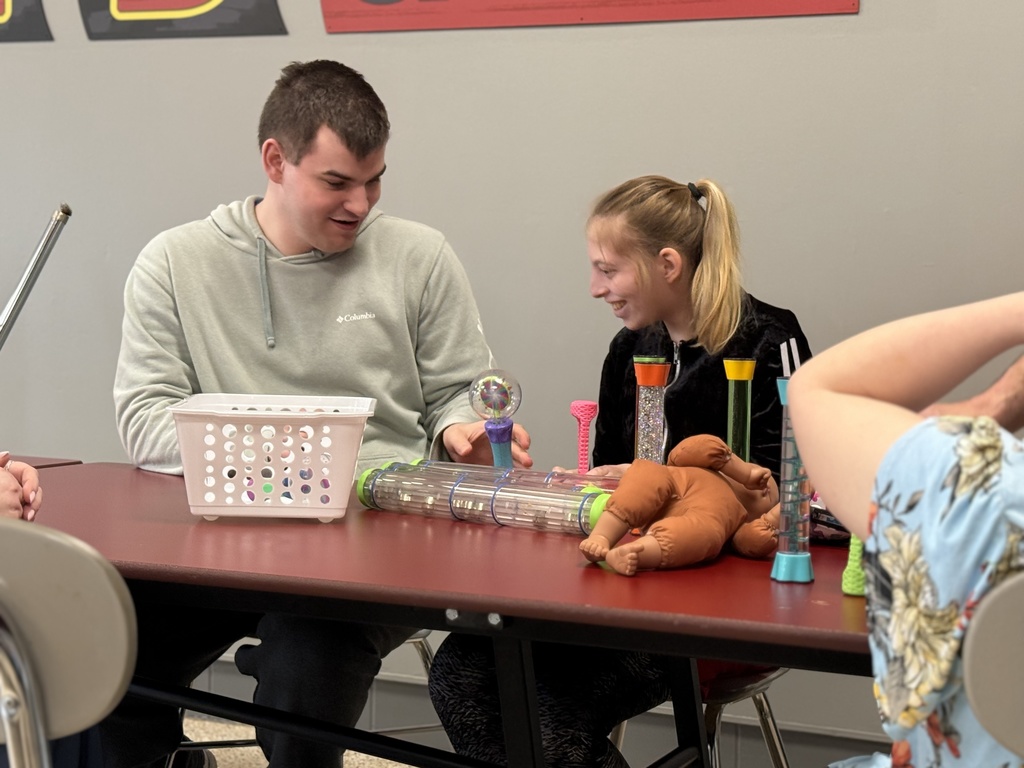 A staff member and a student sit together at a table, smiling and interacting while exploring colorful sensory objects  and toys.