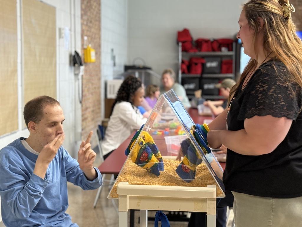 A staff member supports a student at a sensory table with bins and textured materials.