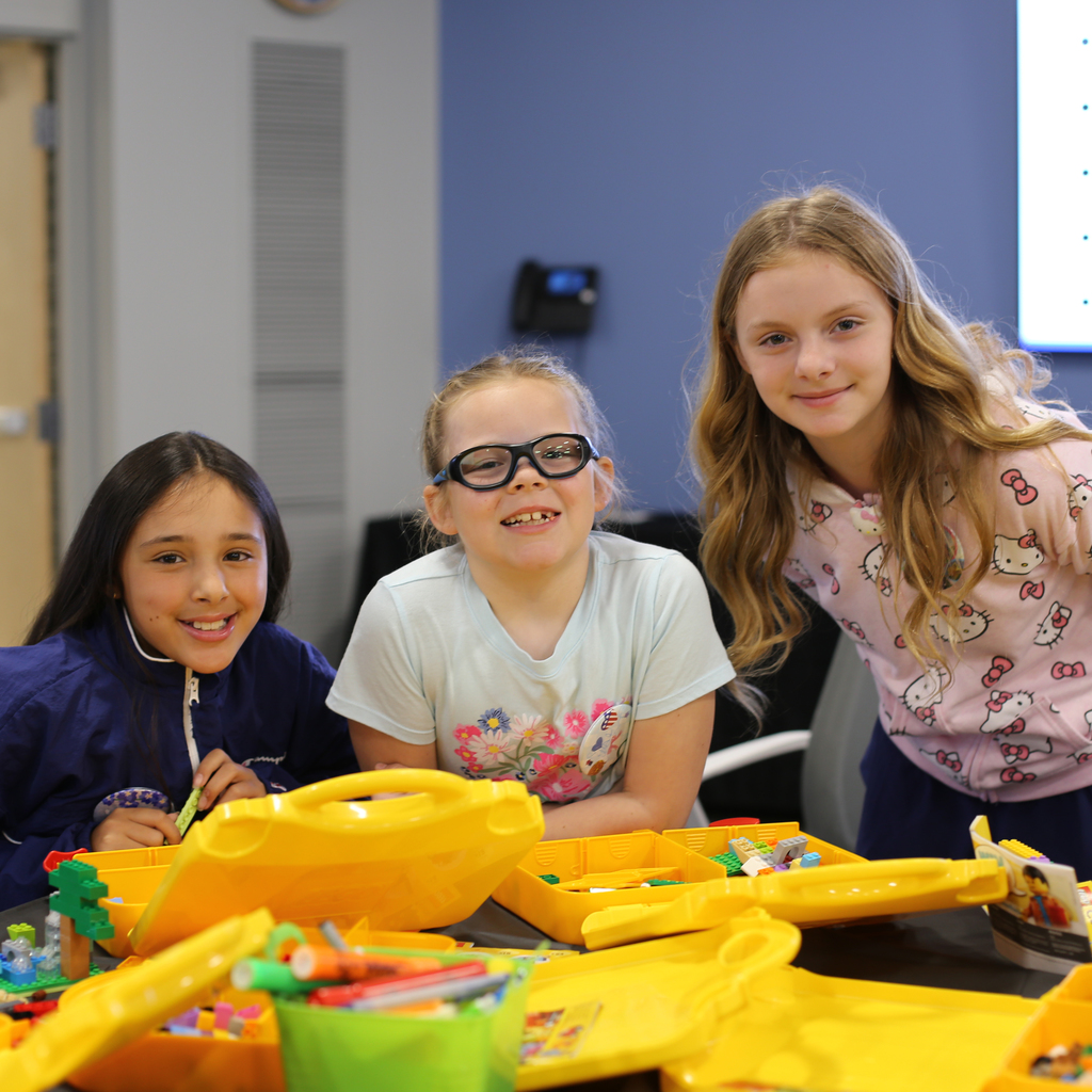 Three students smile at the camera as they sit at a table building with LEGOs