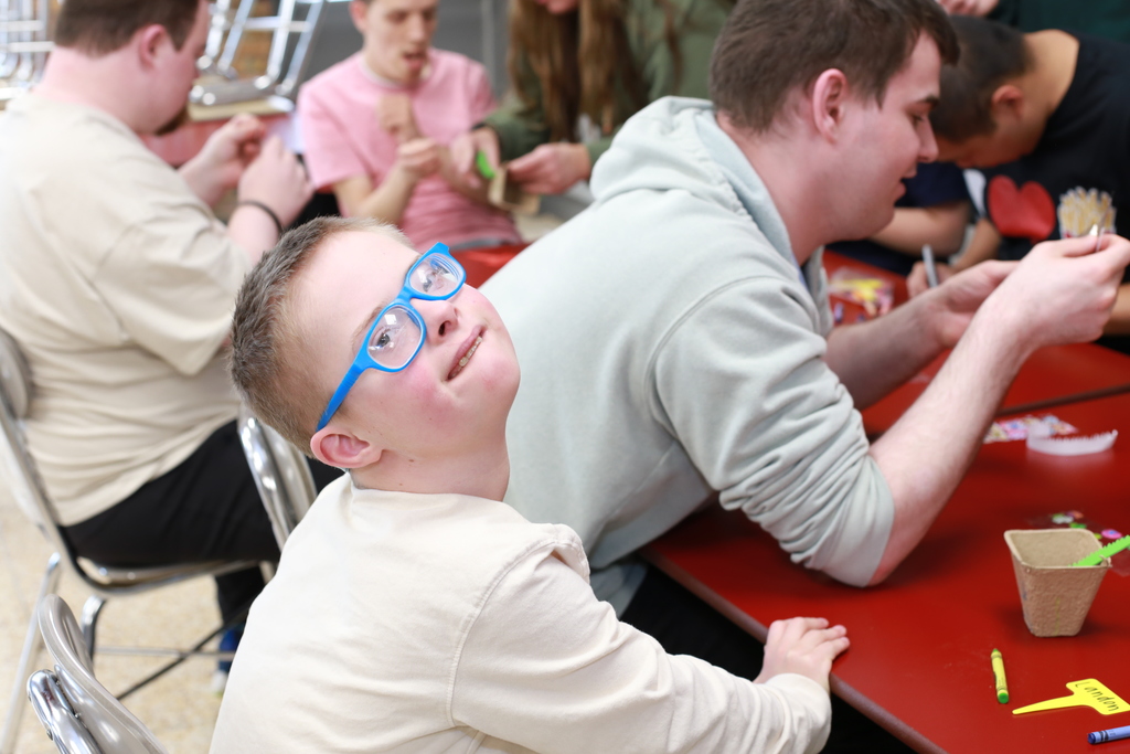A student wearing bright blue glasses looks toward the camera while classmates decorate planters and plant flower seeds during Garden Club.