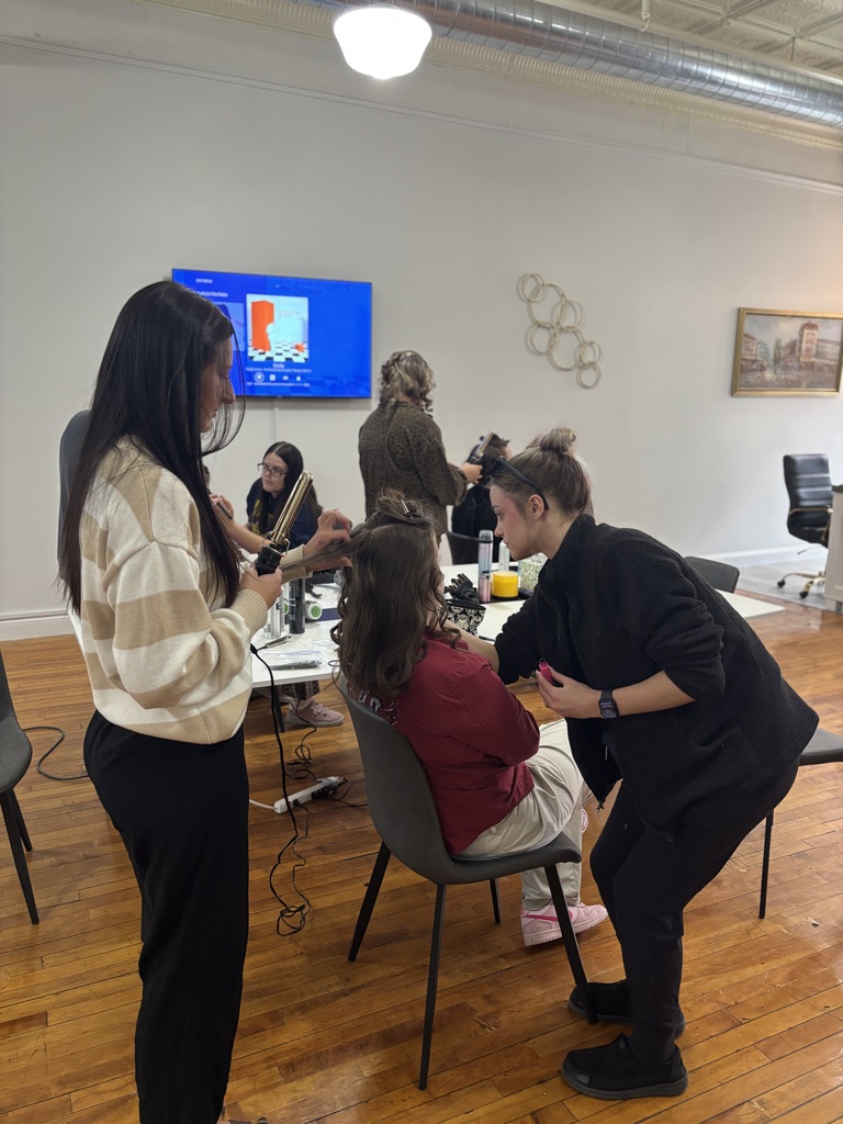Two TEC students apply makeup and curl hair to a seated student while others work in the background as part of the Prom Experience event.
