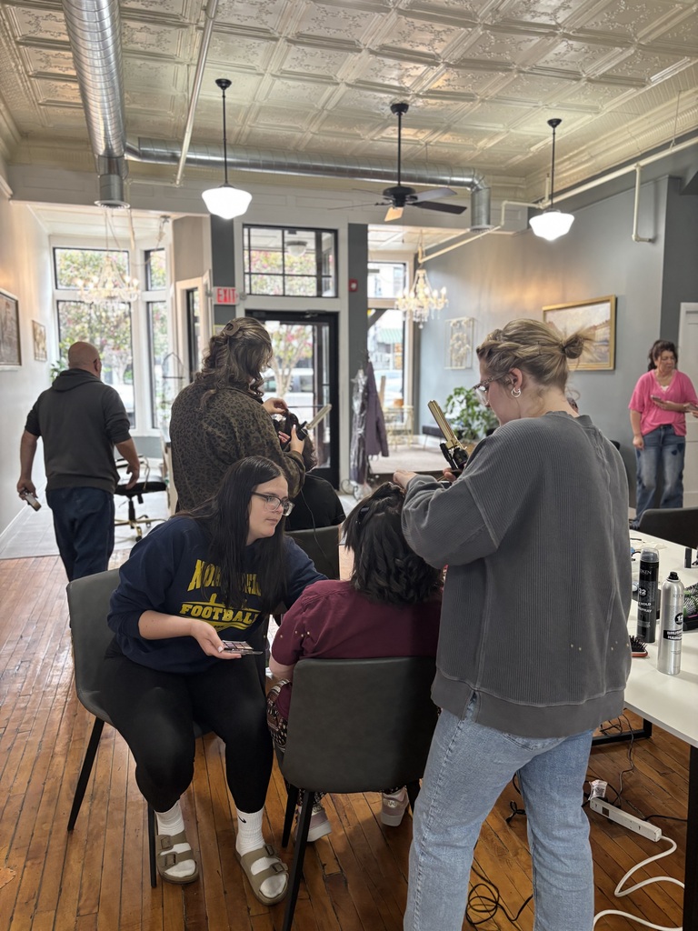 Two TEC students apply makeup and curl hair to a seated student while others work in the background as part of the Prom Experience event.