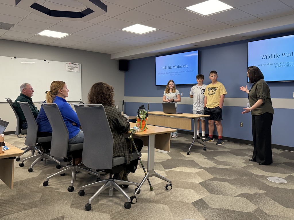 Three students stand in front of two large monitors facing a room of panelists
