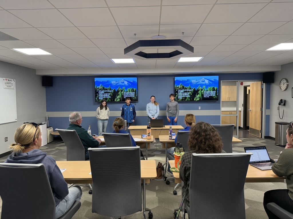 Three students stand in front of two large monitors facing a room of panelists