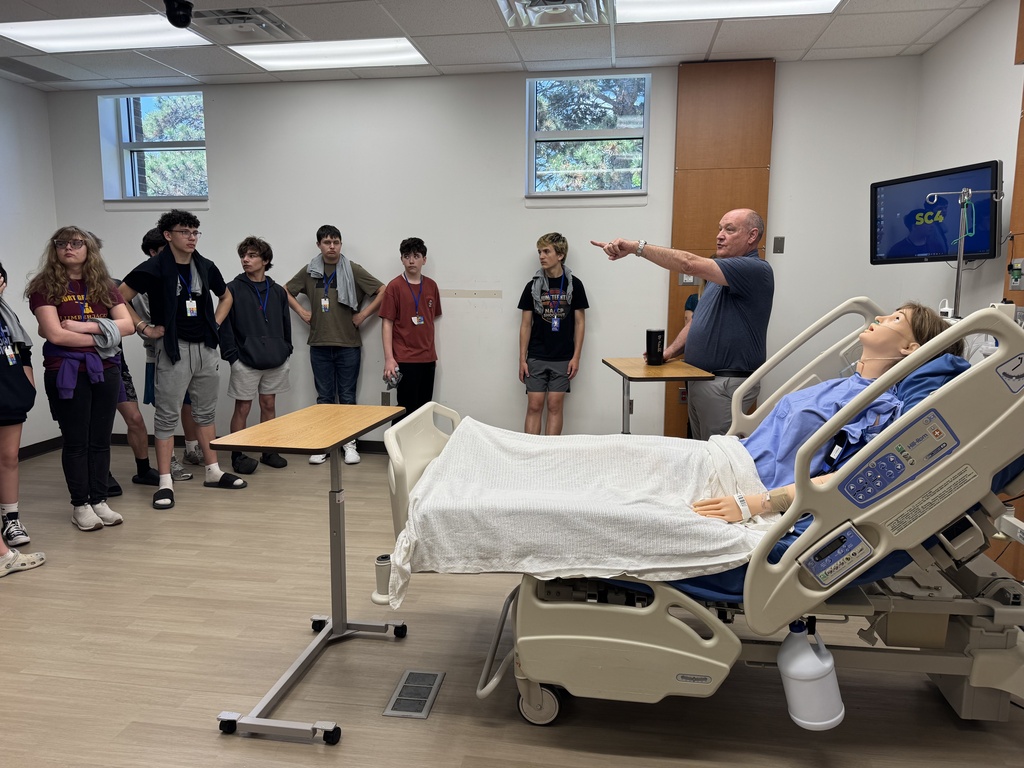 An SC4 standing next to a medical dummy in an hospital bed points to a screen as they talk to a group of eighth graders during the recent college tour.