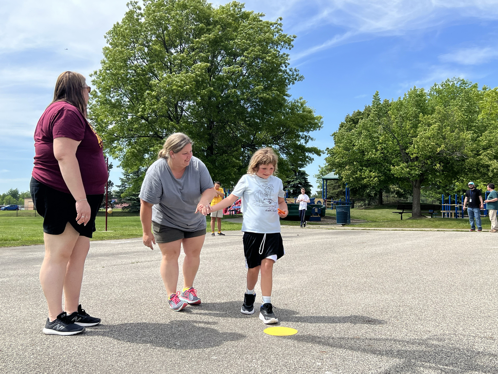 A staff member holds a student's hands as they walk toward a yellow dot on the ground. Another staff member watches.