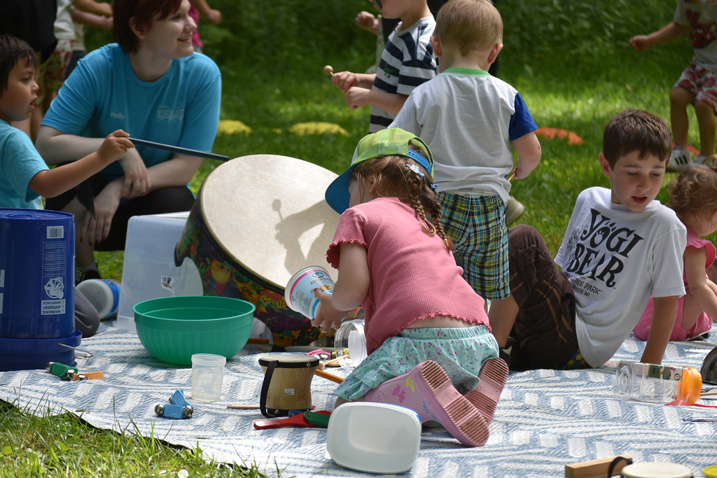 An adult leads children sitting on a blanket in the grass through a musical activity.