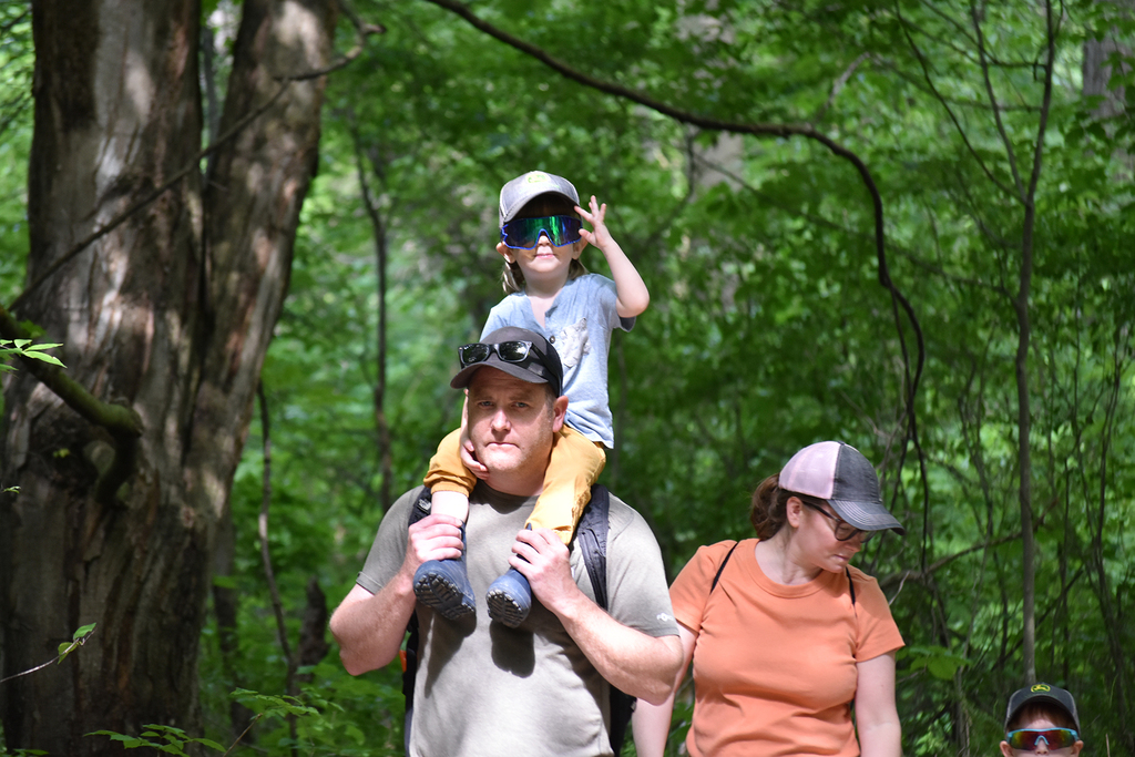 A family walks the trails at the nature center. A toddler sits on the shoulders of a man.