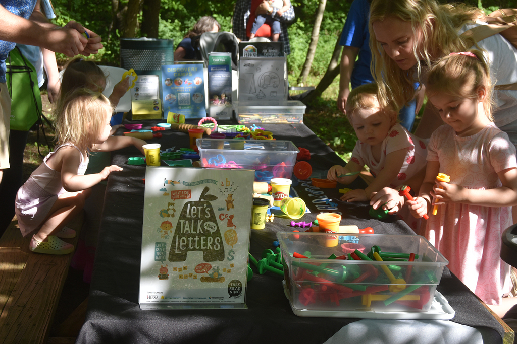 Adults and children stand around a table with Play-Doh and cutout and rolling tools. A sign reads, "Let's talk about letters." 