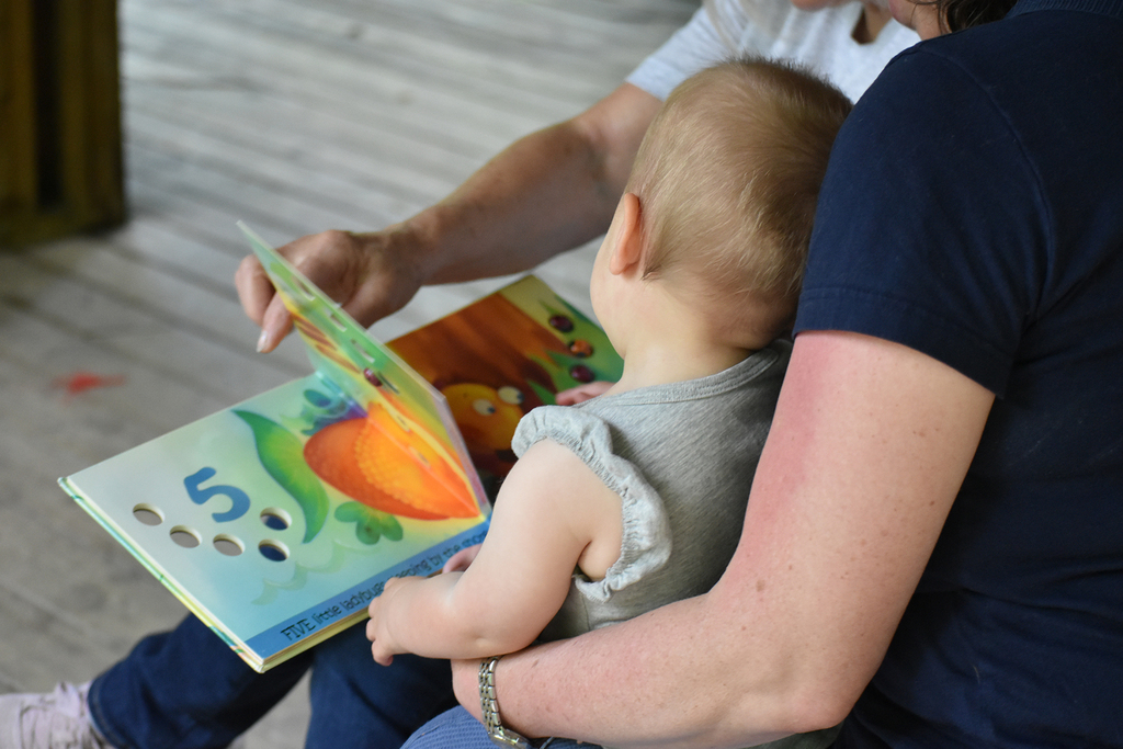 Back view of a toddler sitting on an adult's lap while another adult turns the pages of a board book.