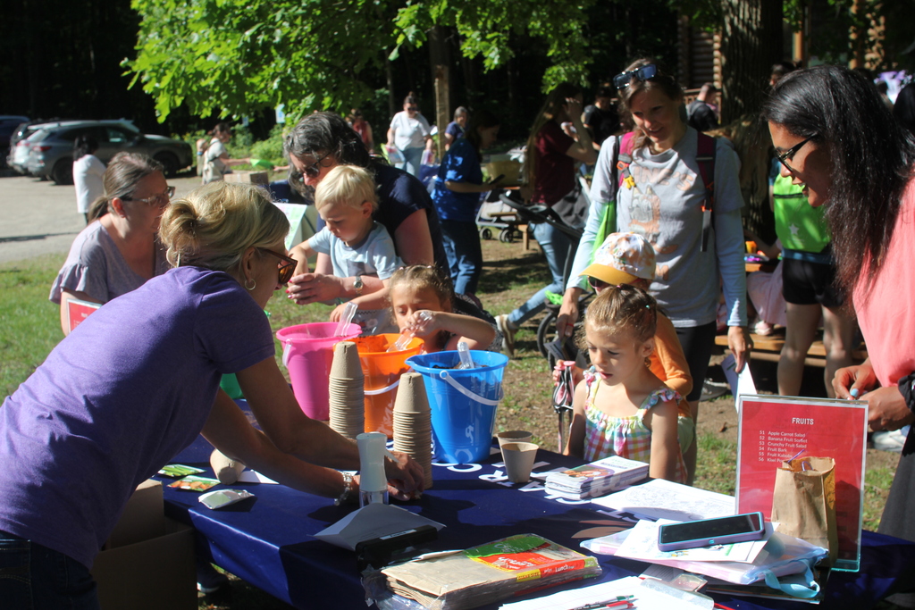 Adults and young children stand around a table participating in a seed planting activity. Groups of people can be seen in the background.