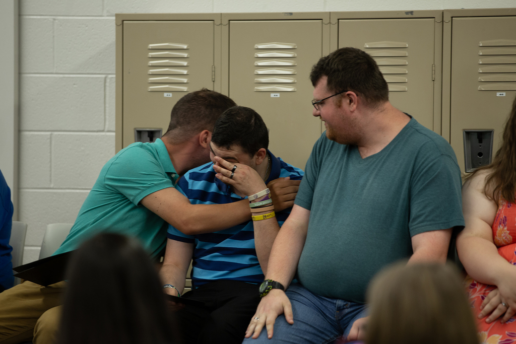 A Next Step student hugs another student who is wiping tear from his eyes, as a third student, smiling, looks on