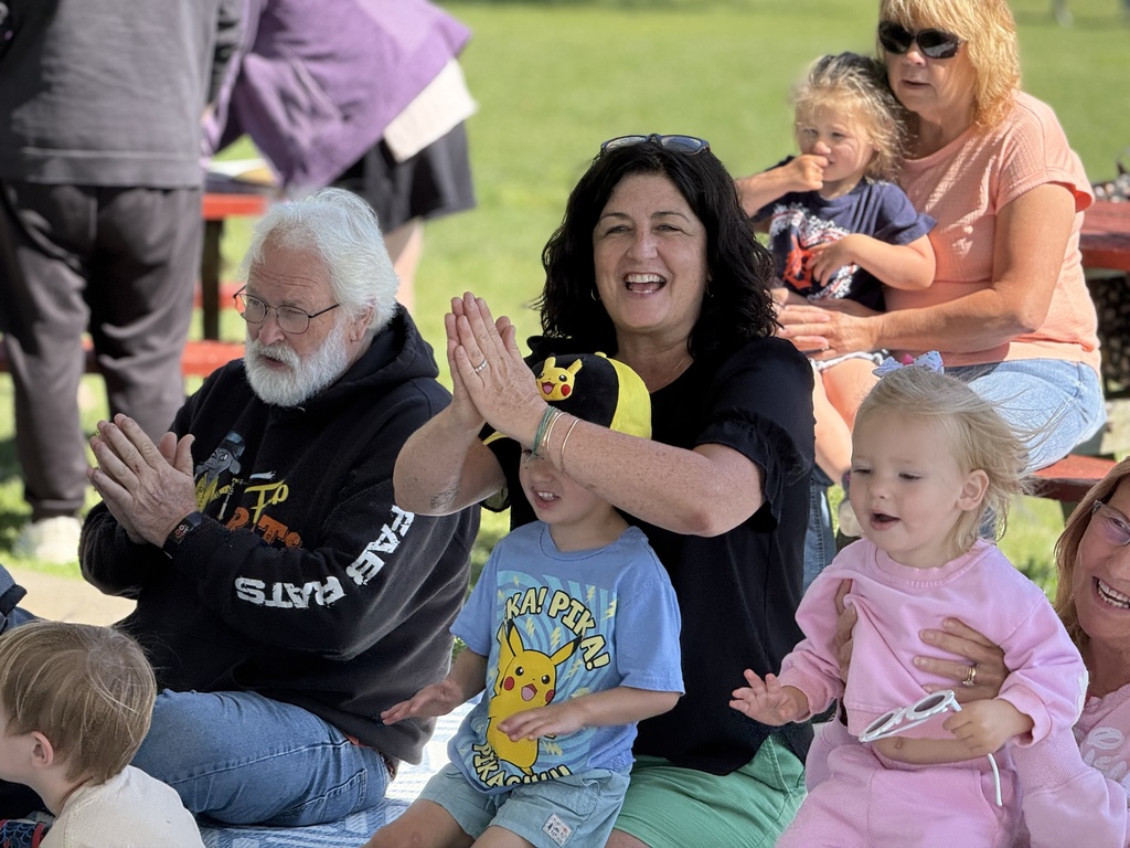 Adults and children sit on the group clapping their hands 