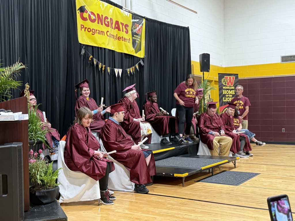Woodland's nine completers wearing graduation caps and gowns sit in chairs on a dais in the school gym with a banner in the background reading, "COngrats Program Completers 