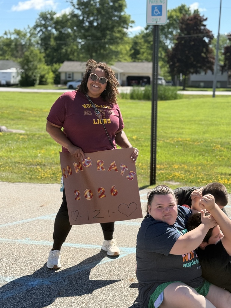 A Woodland staff member standing in front of the school wearing a school shirt and sunglasses holds a sign reading, "Congrats 2026"
