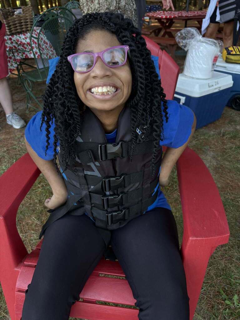 A student wearing glasses and a life jacket sitting in a red Adirondack chair smiles at the camera 