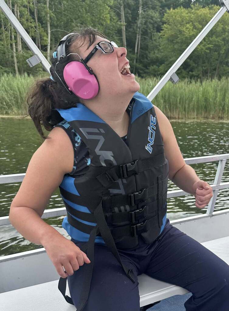 A Woodland student wearing ear protection and a life jacket sits on a pontoon boat and smiles exuberantly