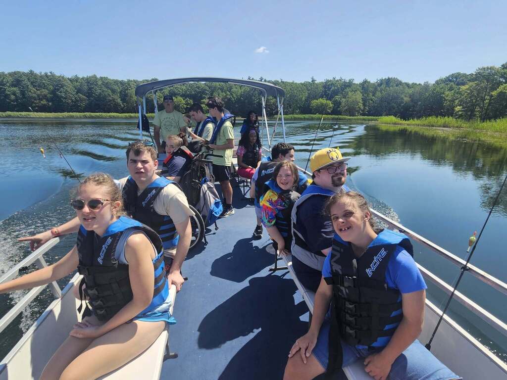 Woodland students and staff wearing life jackets sit on a pontoon boat on the water. Some hold fishing rods