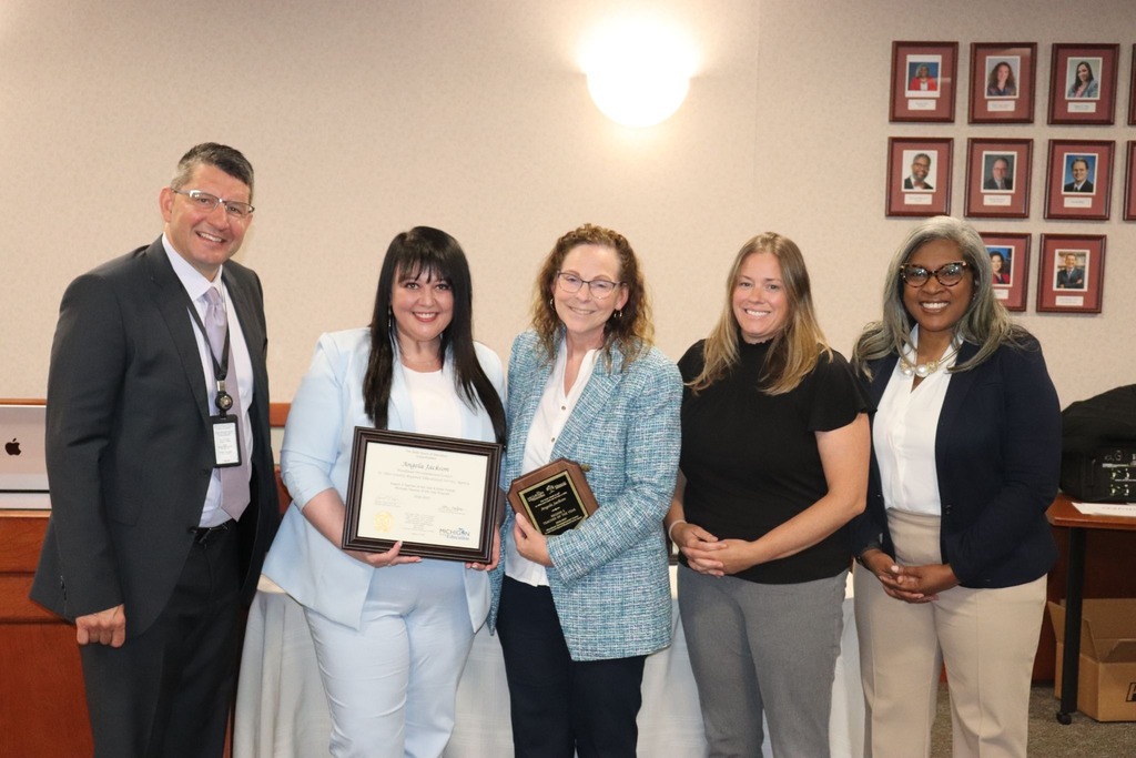 Five people stand together at the Michigan State Board of Education meeting. Woodland teacher Angela Jackson, wearing a light blue suit, holds her Regional Teacher of the Year certificate. 