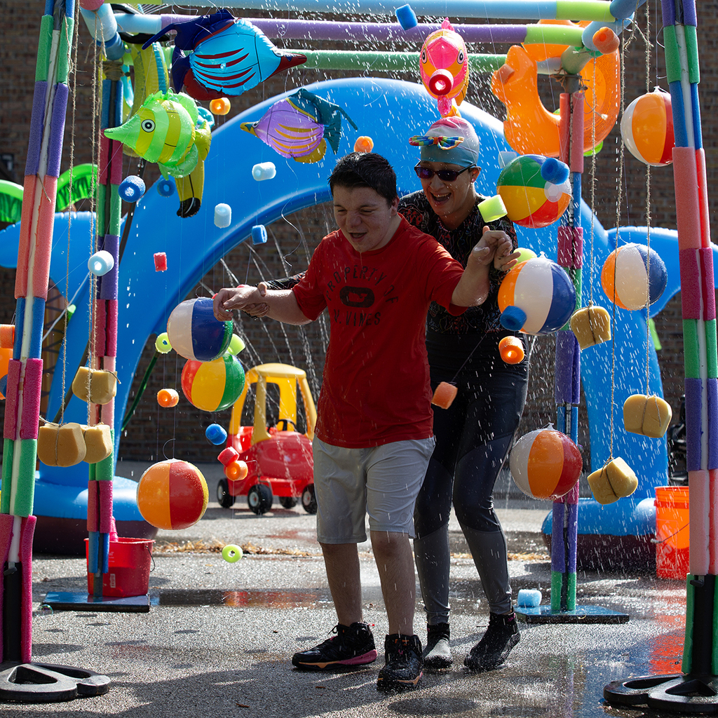 A Woodland staff members holds on to a student as they walk underneath a colorful sprinkler feature