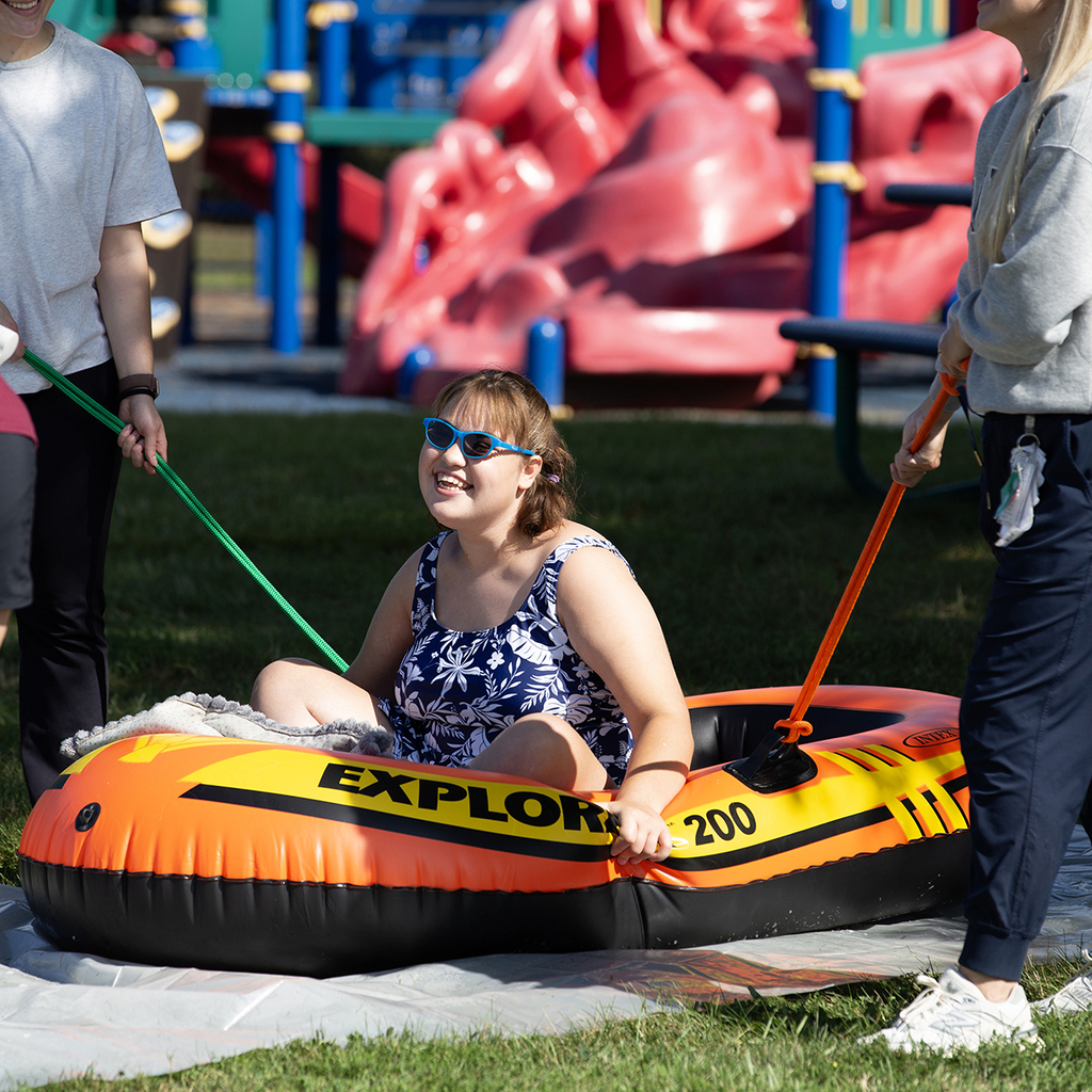 A smiling Woodland student wearing sunglasses and a bathing suit sits in an inflatable raft being pulled by two staff members