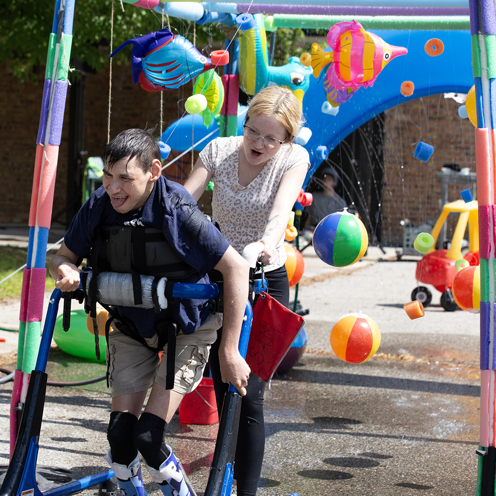 A Woodland staff member pushes a students in a walker under a sprinkler system