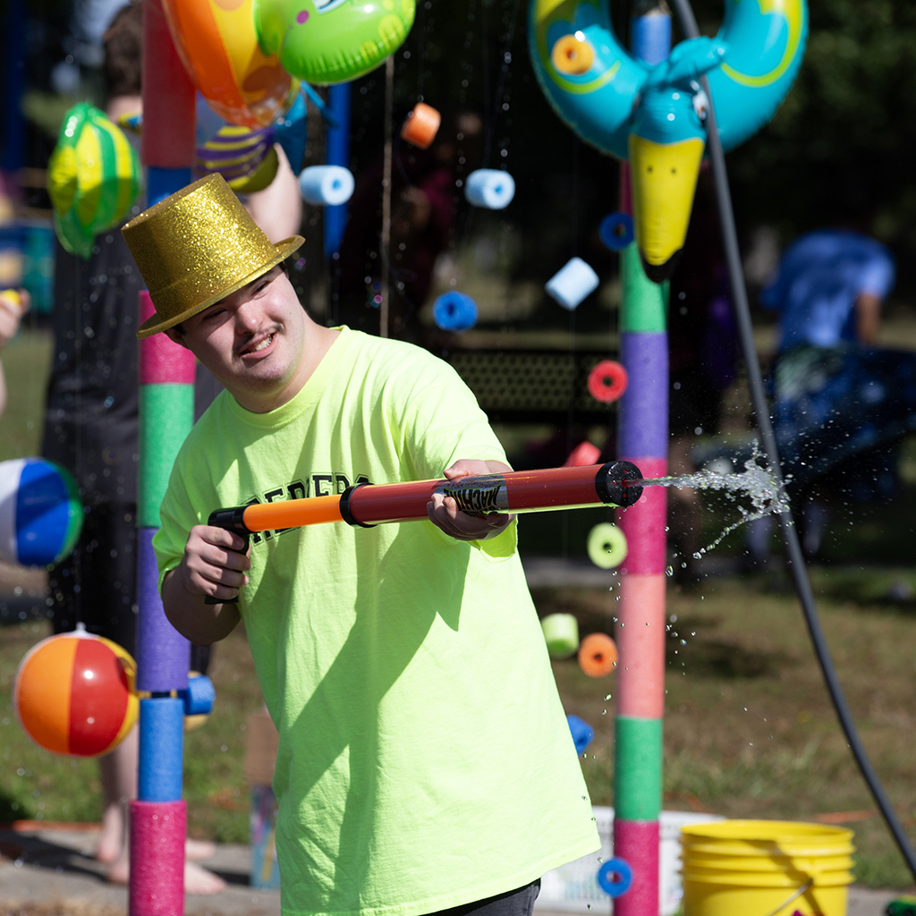 A Woodland student wearing a bright green shirt and sparkly gold top hat, holds a water gun spraying water out the end