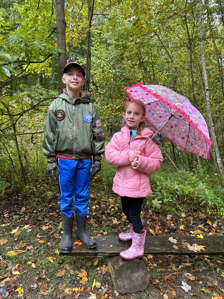 Two elementary age children wearing hats, gloves, and rain boots stand on the nature trail. One holds an umbrella. colorful leaves litter the ground.
