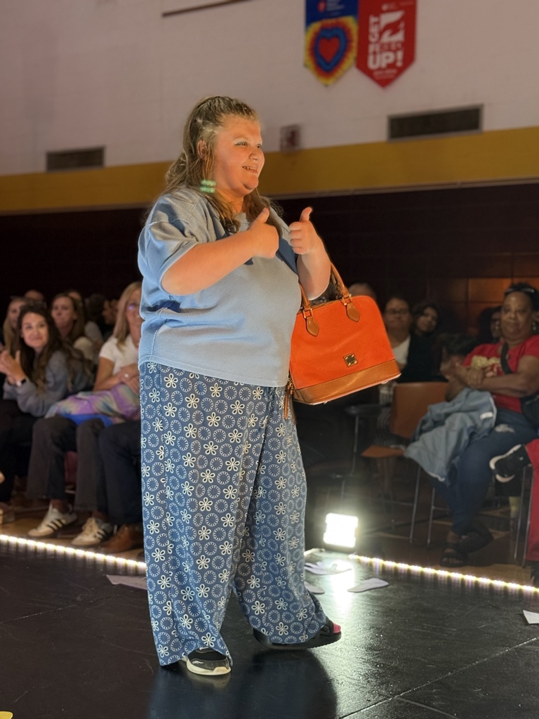 A Woodland student wearing blue floral patterned pants, blue shirt, and holding an orange purse gives two thumbs up to the audience