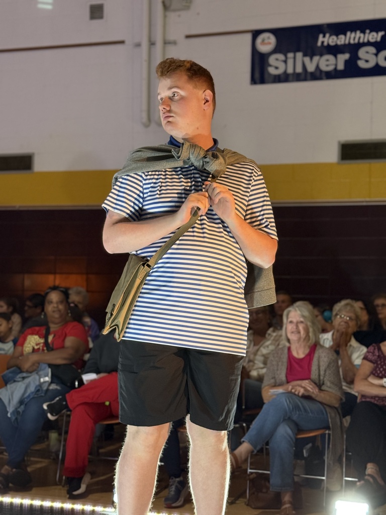 A Woodland student poses at the end of a raised runway. He wears a striped t-shirt with matching shorts, crossbody bag, and jacket tied around his shoulders