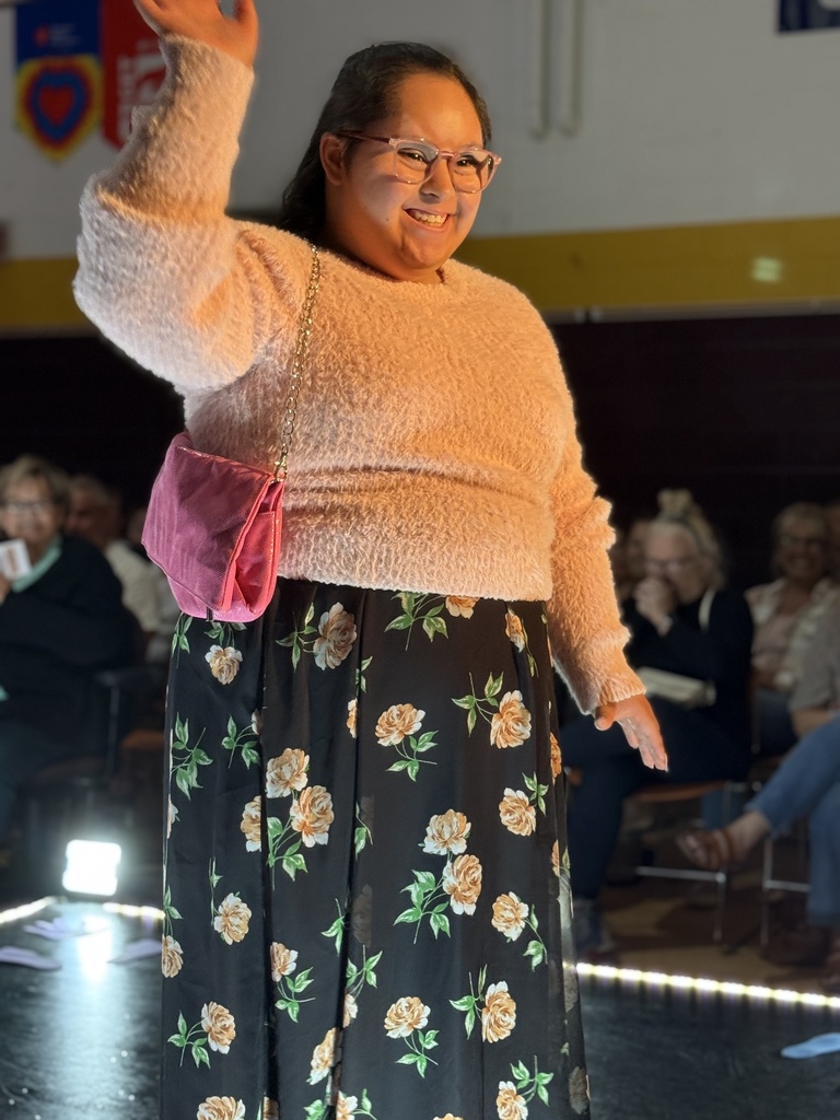 A Woodland student smiles and waves as she walks down the runway. She is wearing a floral long skirt and matching fuzzy sweater