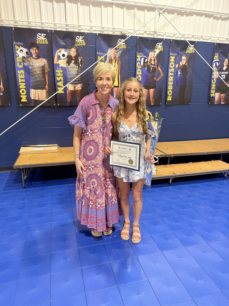 Sacred Heart Academy award recipient smiles with the representative of the school while holding a certificate and flowers.
