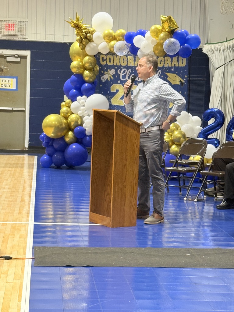 speaker addresses families and students from a podium during the 8th grade Awards Breakfast, with graduation decorations in the background.