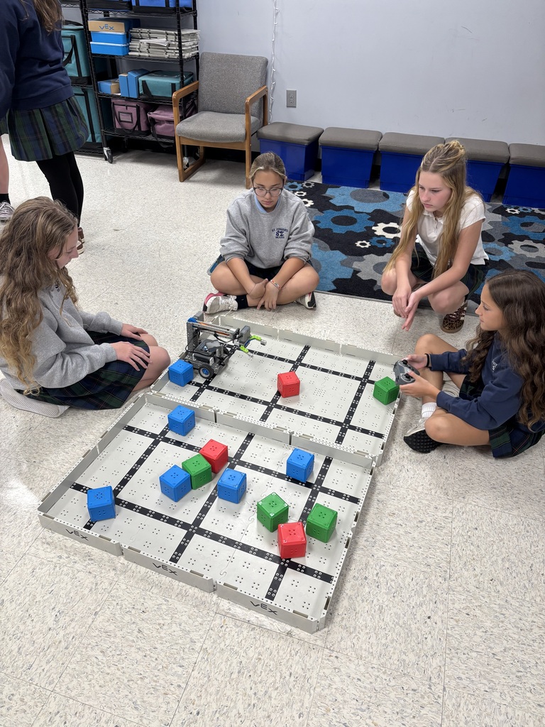 A group of 7th grade students gather around a VEX robotics field, watching and controlling robots as part of a hands on STEM lesson.