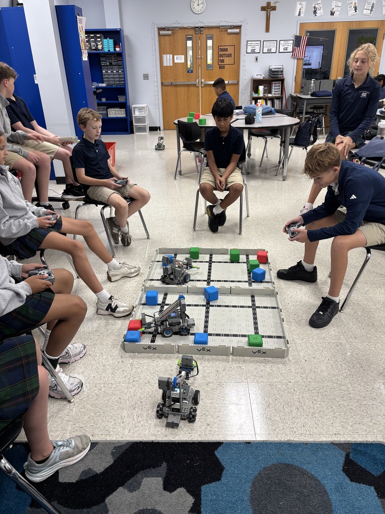 7th grade students sit around a VEX robotics field in the STEM classroom, using controllers to drive robots through a cube challenge.