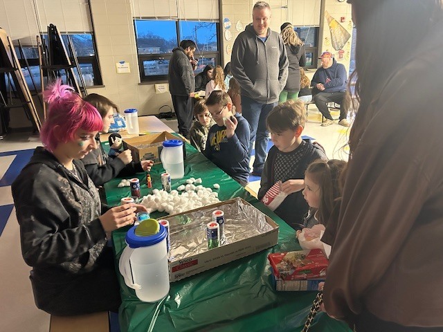 Students and adults sit at a table with science materials.