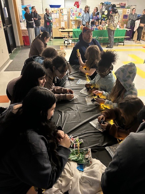 Students sit around a table with some adults and science materials.