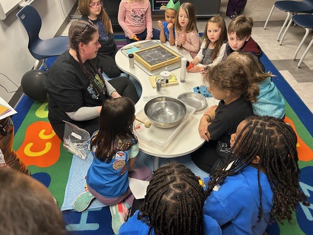 Students sit around a kidney table with an adult and science materials.