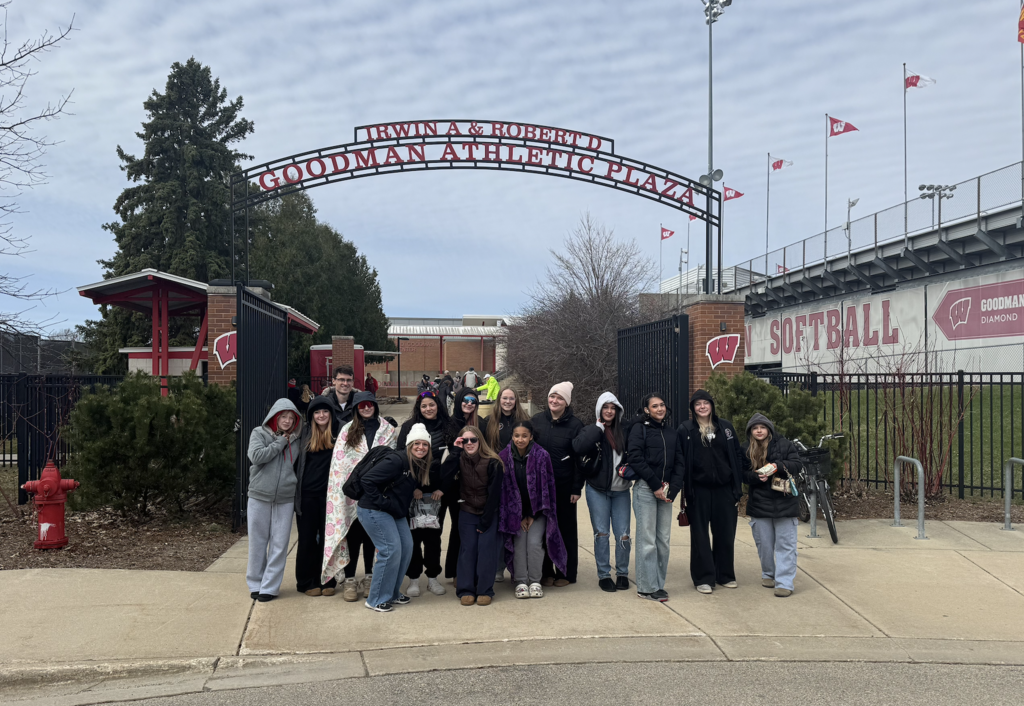 Softball team stand huddled together in front of the Goodman Athletic Plaza sign. 