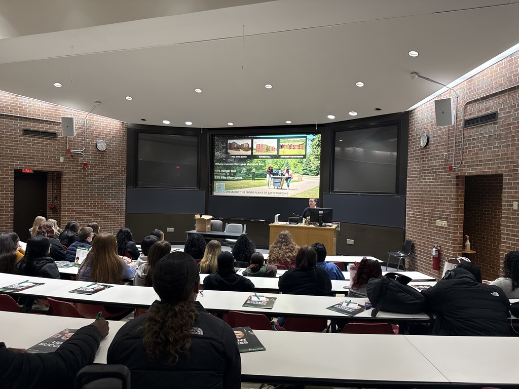 Students sit in a college classroom listening to a speaker with a large screen displaying on the back wall. 