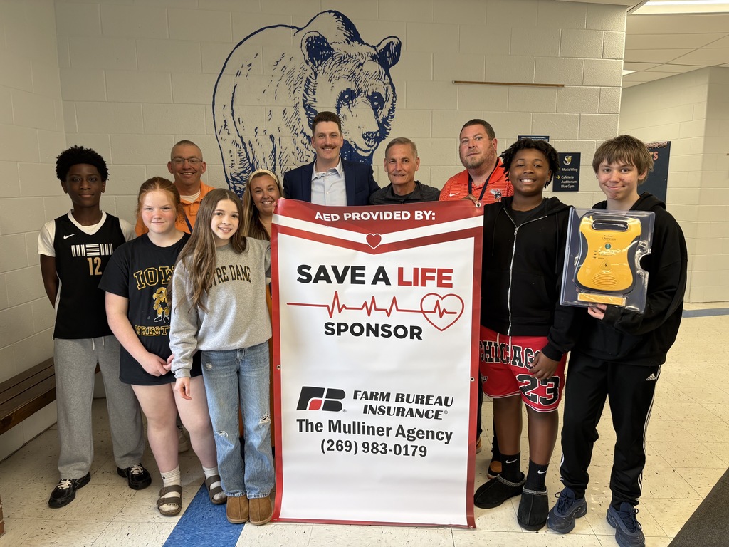 A group of Upton Middle School students and staff stand together in a school hallway in front of a large painted bear mural. They are holding a banner that reads “AED Provided By: Save a Life Sponsor – Farm Bureau Insurance, The Mulliner Agency,” with a phone number listed. One student on the right is holding a yellow automated external defibrillator (AED) device in its case. The group appears to be gathered to recognize the donation of the life-saving equipment to the school.
