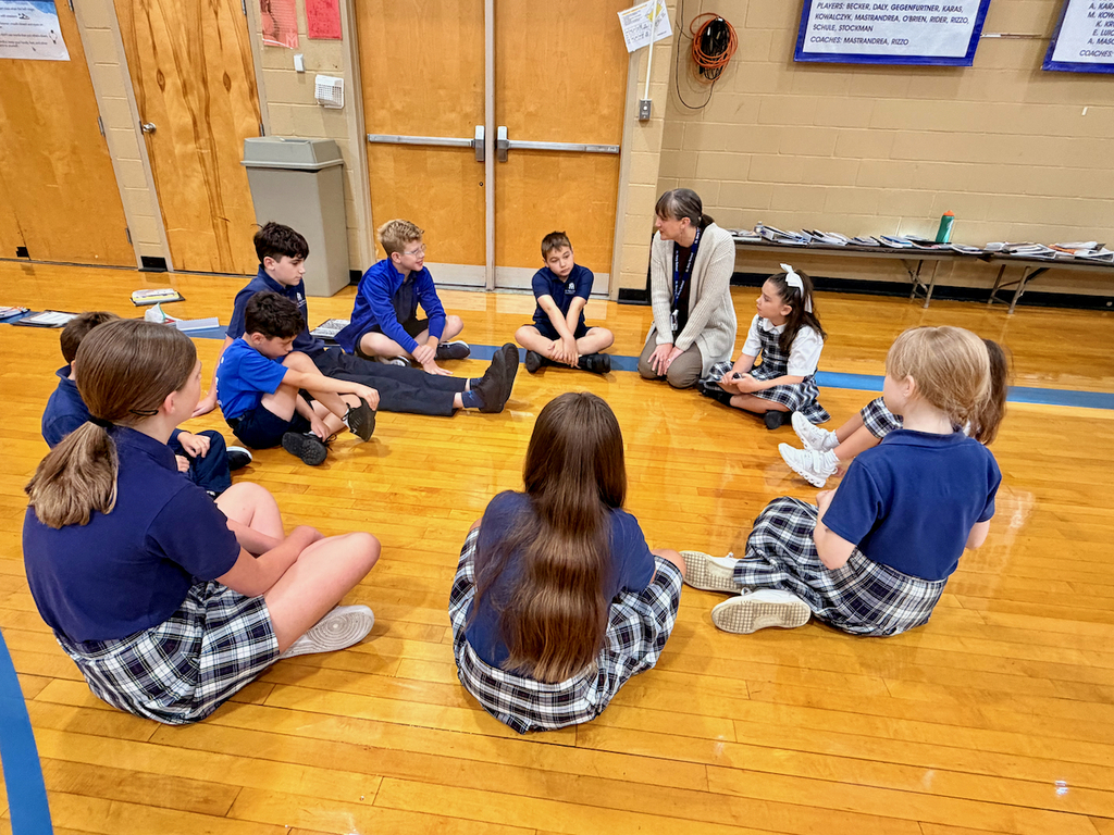 group of students sitting in a cirlce on a gymnasium floor with their teacher