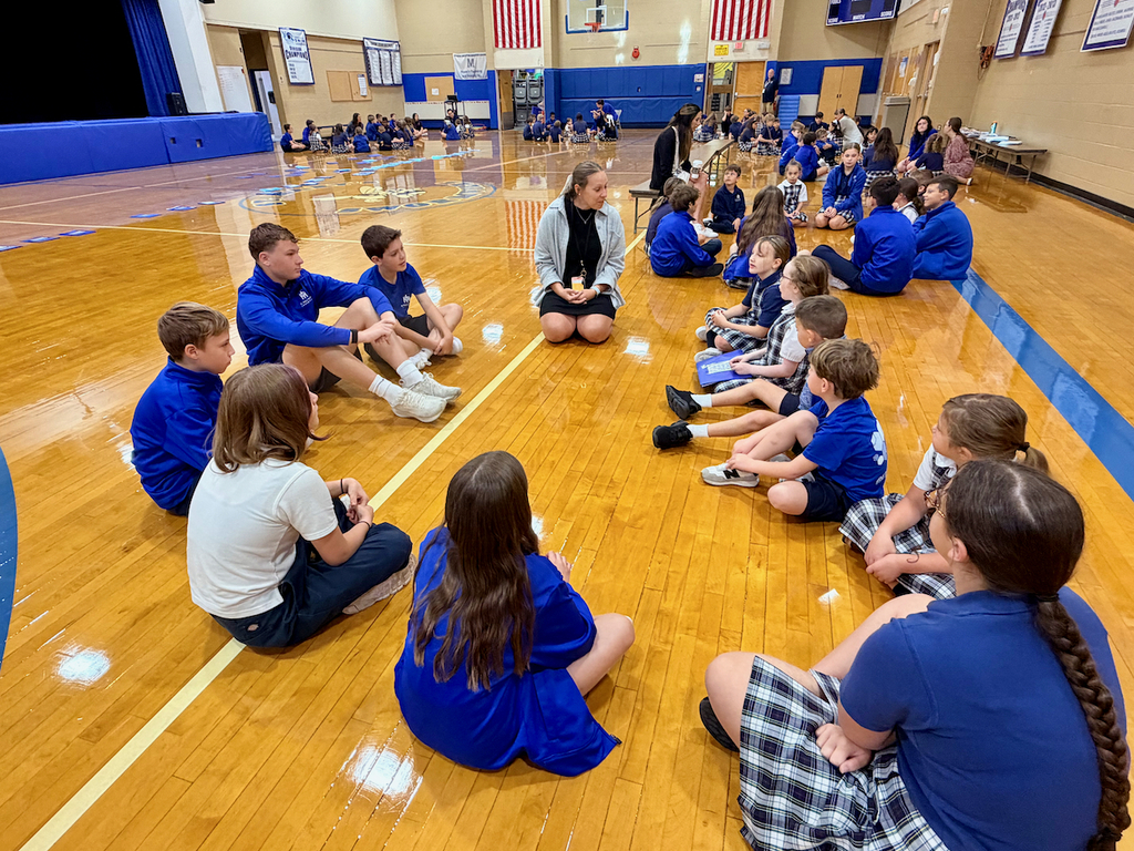 group of students sitting in a cirlce on a gymnasium floor with their teacher