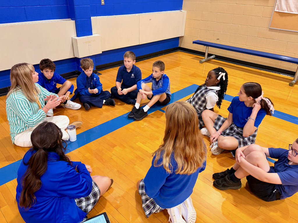 group of students sitting in a cirlce on a gymnasium floor with their teacher