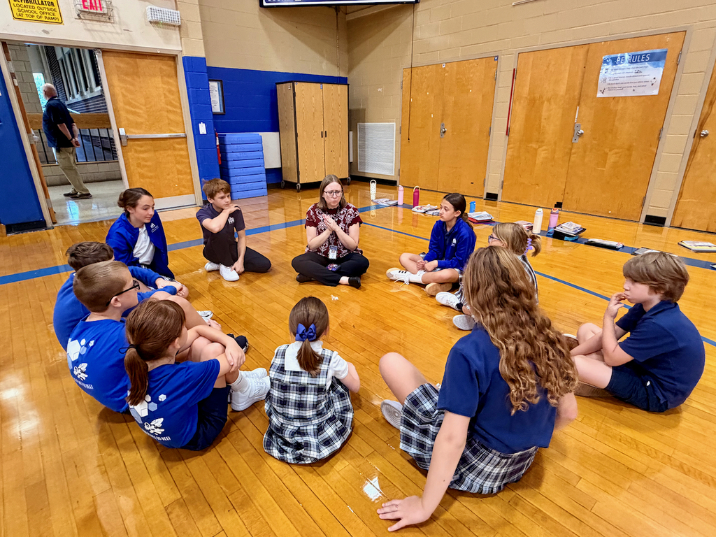 group of students sitting in a cirlce on a gymnasium floor with their teacher