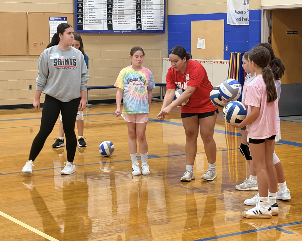 girls learning to serve volleyballs in gym