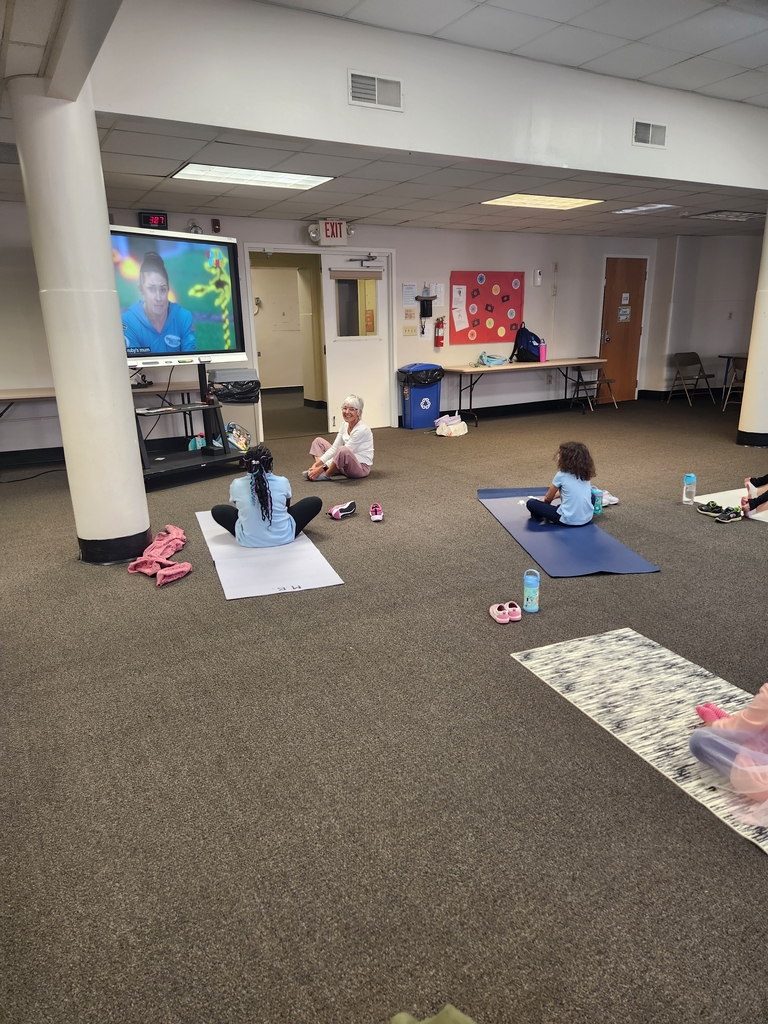 Teacher standing next to smart board which has the yoga up from YouTube. Students sit in front of smartboard copying yoga moves.