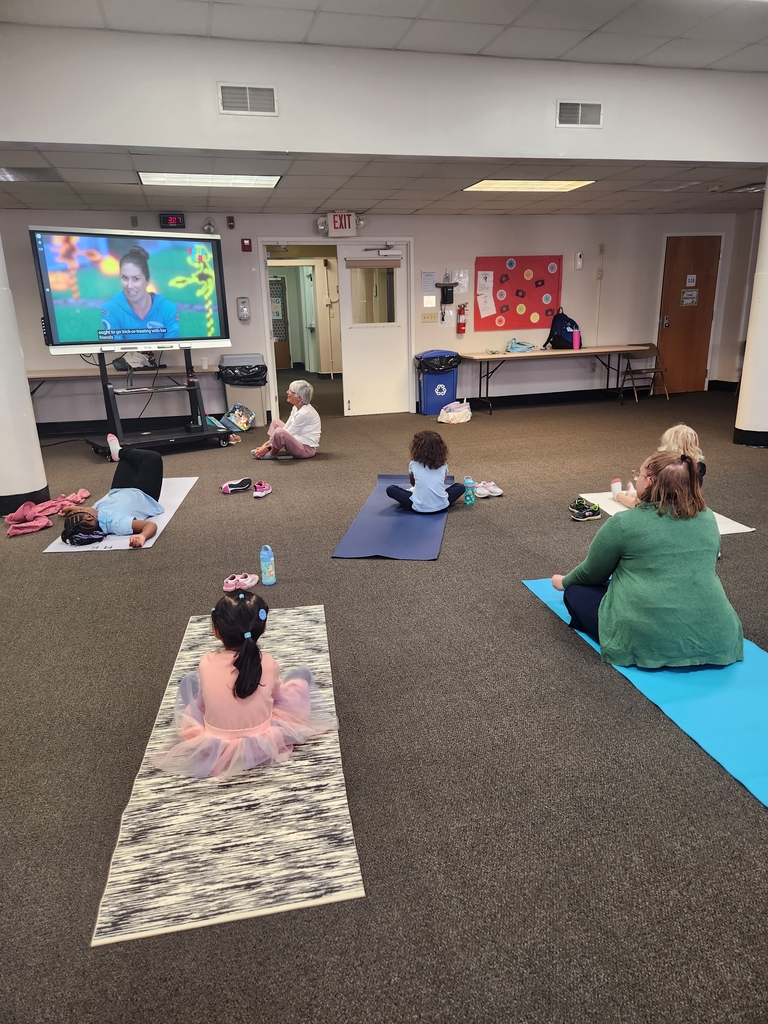 Teacher standing next to smart board which has the yoga up from YouTube. Students sit in front of smartboard copying yoga moves.